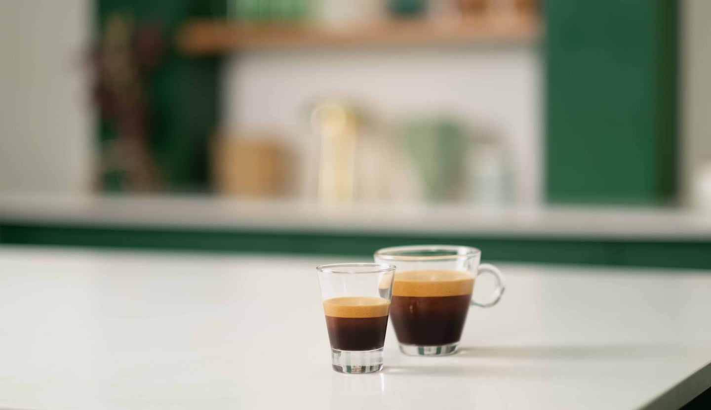 Dvě sklenice stojící na bílém stole - jedna s Starbucks® Ristretto Shot, druhá s Starbucks® Blonde nebo Dark Espresso.