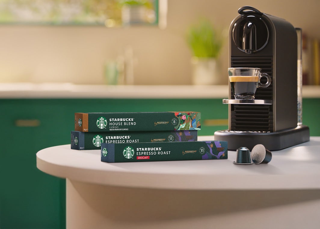 Eine Nespresso Maschine bereitet einen Espresso in einer kleinen Tasse zu, daneben liegen zwei gestapelte Packungen Starbucks